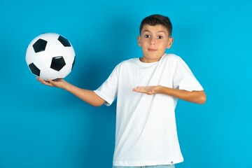 Little hispanic boy wearing white T-shirt holding a football ball pointing aside with both hands...