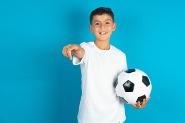 Little hispanic boy wearing white T-shirt holding a football ball Pointing with finger surprised...