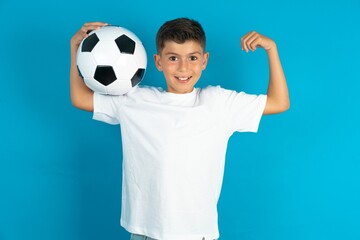 Little hispanic boy wearing white T-shirt holding a football ball showing arms muscles smiling...