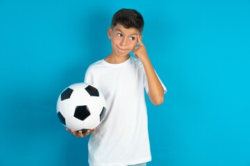Little hispanic boy wearing white T-shirt holding a football ball tries to memorize something, keeps fore finger on temple, reminds information for exam,