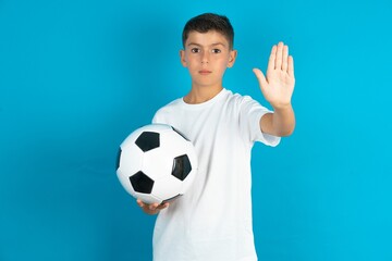 Little hispanic boy wearing white T-shirt holding a football ball doing stop sing with palm of the...