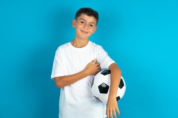 Little hispanic boy wearing white T-shirt holding a football ball happy face smiling with crossed arms looking at the camera. Positive person.