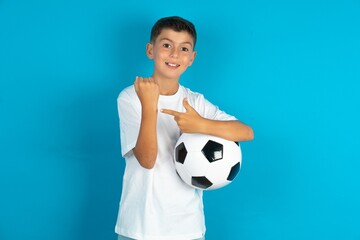 Little hispanic boy wearing white T-shirt holding a football ball In hurry pointing to wrist watch,...
