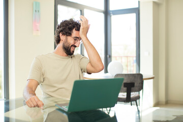 young adult bearded man with a laptop laughing and slapping forehead like saying d’oh! I forgot...