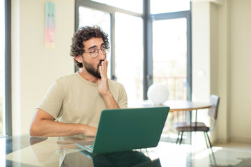 young adult bearded man with a laptop yawning lazily early in the morning, waking and looking...