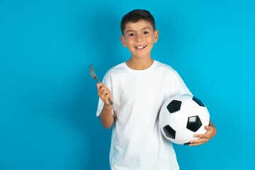 hungry Little hispanic boy wearing white T-shirt holding a football ball holding in hand fork knife...
