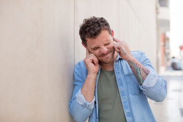 handsome man looking angry, stressed and annoyed, covering both ears to a deafening noise, sound or...