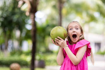 Candid lifestyle portrait of cute little Caucasian girl nine years old with blonde hair in stylish pink dress holding hands fresh coconut with surprised face when walking outdoor in tropical garden