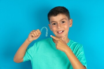 Little hispanic boy wearing green T-shirt  holding an invisible aligner and pointing at it. Dental healthcare and confidence concept.