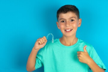 Little hispanic boy wearing green T-shirt holding an invisible braces aligner and rising thumb up,...