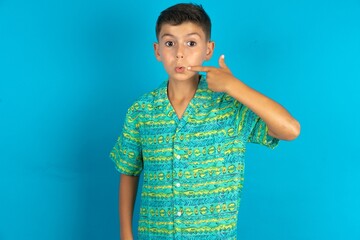 Charming Little hispanic boy wearing green aztec shirt pointing on pout lips with forefinger,...