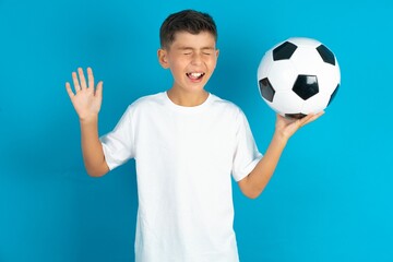 Emotive Little hispanic boy wearing white T-shirt holding a soccer ball laughs loudly, hears funny...