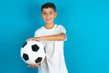 Little hispanic boy wearing white T-shirt holding a soccer ball gesturing with hands showing big and large size sign, measure symbol. Smiling looking at the camera.