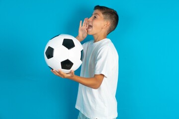 Little hispanic boy wearing white T-shirt holding a soccer ball shouting and screaming loud to side with hand on mouth. Communication concept.