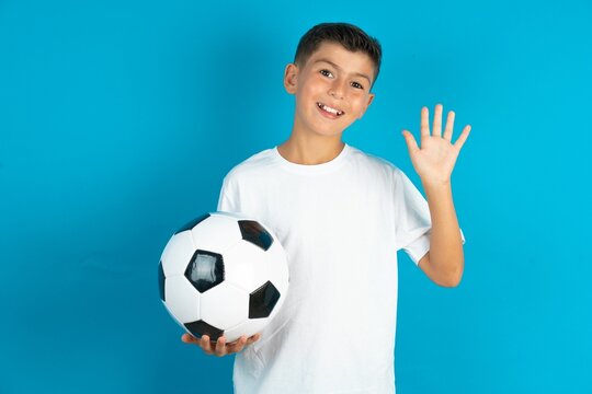 Little hispanic boy wearing white T-shirt holding a soccer ball Waiving saying hello happy and smiling, friendly welcome gesture.