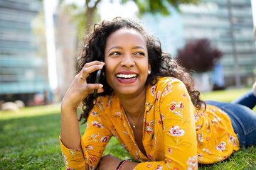 Happy young black woman lying on grass and talking on mobile phone outside