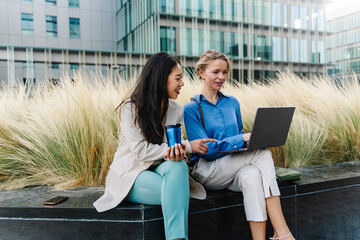 Women coworkers working on laptop sitting outside business corporate office building