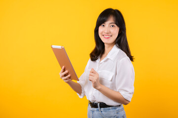 young Asian woman wearing white shirt smiles brightly while holding digital tablet against yellow background, represents a confident company worker embracing modern technology concepts.