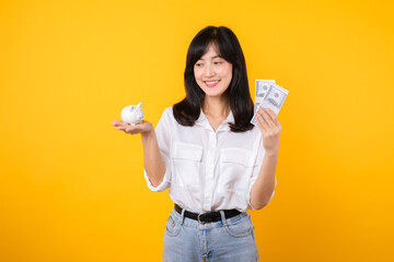 Portrait beautiful young business asian woman wearing white shirt and denim jean with a lot of cash money and piggy bank isolated on yellow background. Wealth money saving, finance investment concept.