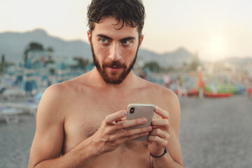 Pensive Young Man with Smartphone on Beach - A young Caucasian man in swimwear holds a smartphone, gazing off-camera with a questioning look, with blurred beach in the background.