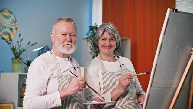 hobies and entertainment, portrait of elderly talented pensioners with brushes in their hands paint a picture with paints on canvas at home, smile and look at camera