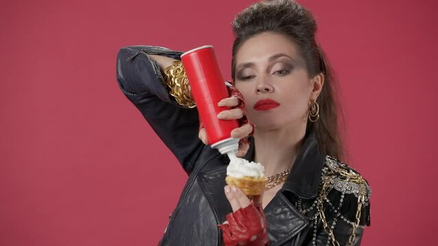 A woman applies whipped cream from an aerosol can to a cupcake, then squeezes the whipped cream into her mouth, winks. Portrait of a woman on a red background close up. HDR BT2020 HLG Material.