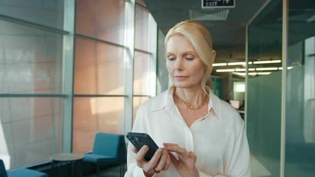 Mature businesswoman 50s walking at office corridor holding smartphone, looking attentively at mobile phone screen, scrolling news, reading message, thinking through decision concerned face expression