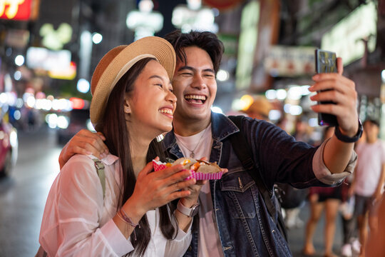 Asian Couple Tourist Backpacker Doing Selfie And Recording A Video Blog While Eating Food From Street Stall In Night Market With Crowd Of People At Yaowarat Road, Bangkok