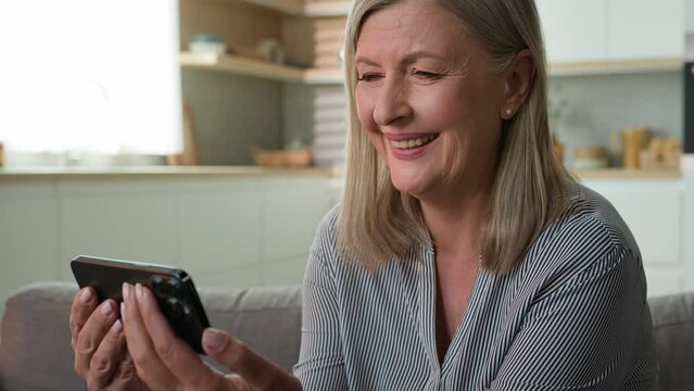Smiling elderly caucasian woman looking mobile phone screen laughing watching funny video on internet browsing photo having fun online reading good news on social network using smartphone at home sofa