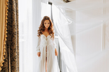 a beautiful girl in a white open dress touches poses standing in a white curtain on the background of a balcony window