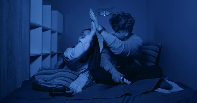 Joyful couple male and female gamers giving high fives after winning team game on console, sitting on bed in evening at home in warm light with joysticks in hands and smiling.
