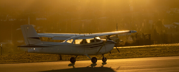 A small Cessna single propeller airplane taxiing down the runway backlit under a yellow sunset sun...