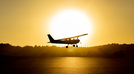 Naklejka na ściany i meble Backlit silhouette of a cessna plane outlined with the sun just behind it flying low with the sea water below illuminated by the golden light of sunset and trees and vegetation in the background
