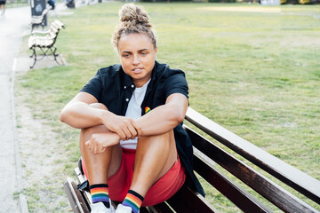 Sad frustrated lesbian woman with LGBTQ rainbow badge on t-shirt sitting on the park bench alone....