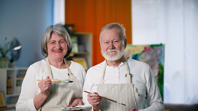 portrait of an old man and woman in aprons with brushes and a palette on background of painted picture, happy woman jokes and paints her husband nose, smile and look at camera