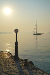 Sailing yacht in morning sunlight in the Aegean sea, with a lantern on the mole of the port of Spetses  