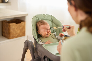 Mother trying spoon feeding her crying baby boy in the kitchen