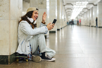 Happy teenage boy waving hand to his friend on smartphone screen while sitting on skateboard in subway tunnel and communicating in video chat