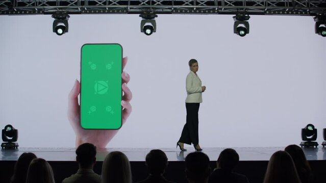 A woman presenter is standing on a well-lit stage in front of a large LED screen showing a hand holding a generic smartphone with green screen. Technology, application or product launch presentation 