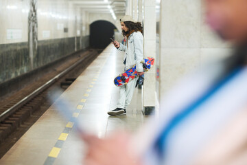 Focus on teenage guy with skateboard texting in smartphone while standing by marble column in front of railway at subway station