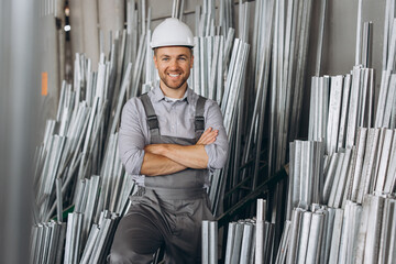 Happy bearded male factory worker in special uniform and white hard hat holding aluminum frame at production of metal plastic windows and doors