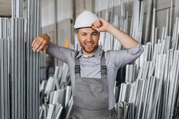 Happy bearded male factory worker in special uniform and white hard hat holding aluminum frame at...