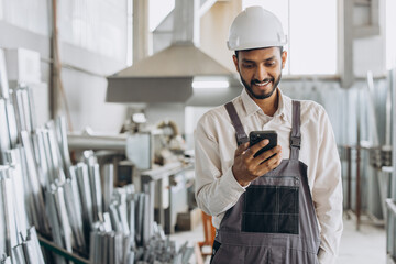 Happy bearded Indian male factory worker in special uniform and white hard hat holding the phone and having a dialogue at the production of metal-plastic windows and doors