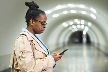 Side view of young black woman looking at smartphone screen while standing in front of camera against subway tunnel in metro