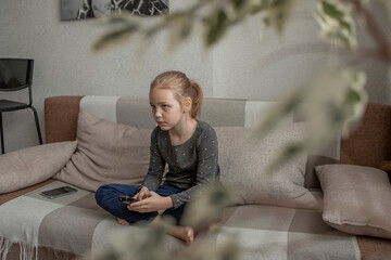 A cute little girl is sitting on the sofa at home in the living room and switches channels on the TV with a remote control. Watching children's shows or videos. Free time after school