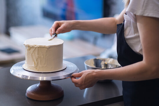 Professional Confectioner Finishing Pattern With Knife On White Mastic Of Cake Using Special High Stand On Kitchen Table Woman In Apron Making Organic Sugar Free Pastry For Client At Home