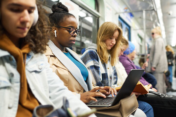 Young serious businesswoman in earphones typing on laptop keyboard while sitting among other passengers traveling by subway train