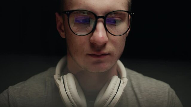 Concentrated young professional programmer in computer glasses with monitors reflection works online late. Serious hacker, IT specialist using laptop computer. Esport player in virtual competition.