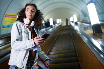 Teenage skateboarder with smartphone standing on moving escalator and looking aside against long...