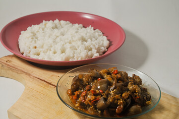 Stir-fried purple eggplant with anchovies and peanuts served in a small plate on a wooden cutting board and a plate of white rice isolated on a white background